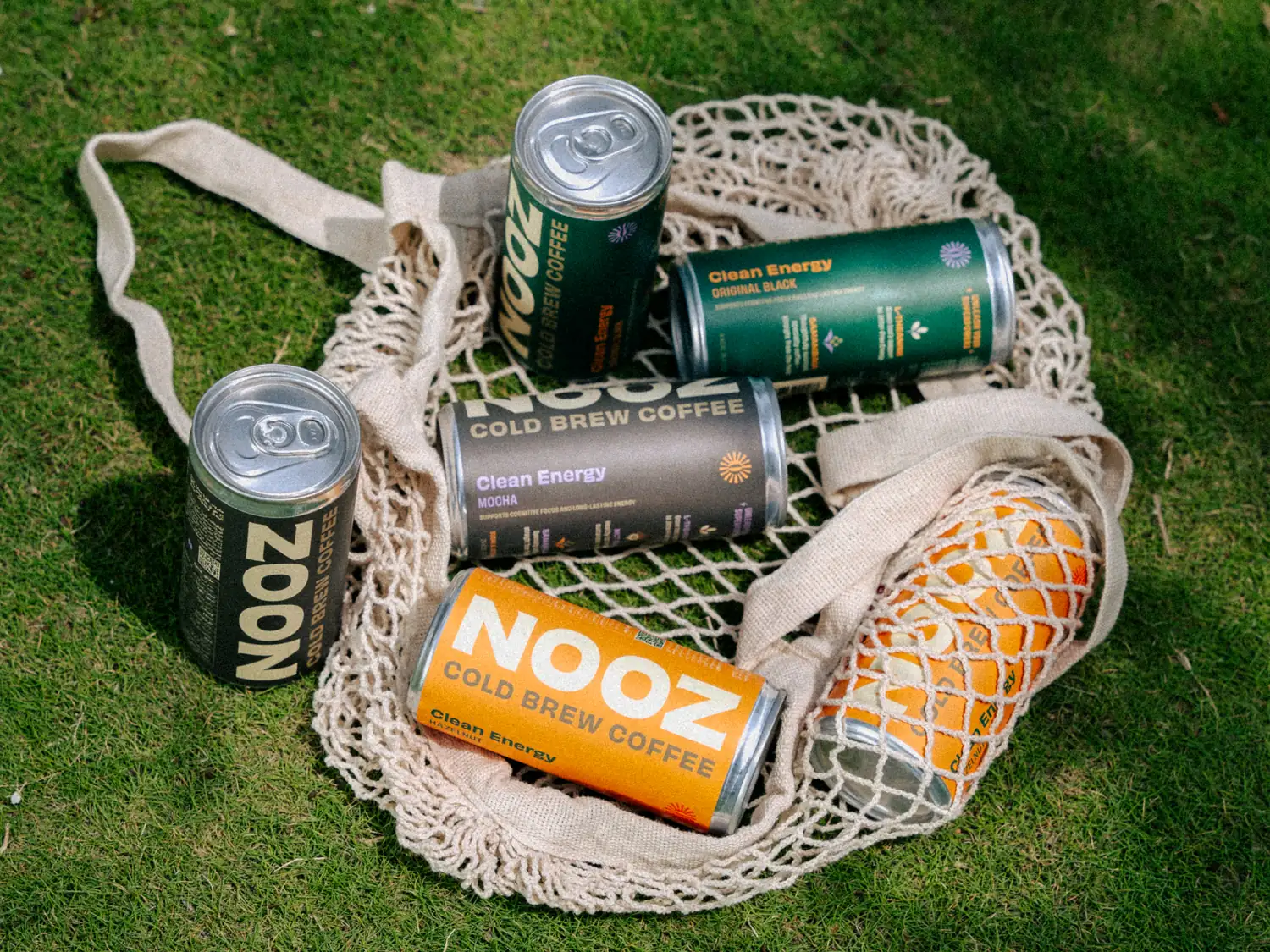 NOOZ cold-brew coffee cans and fresh oranges in a cotton string bag on grass